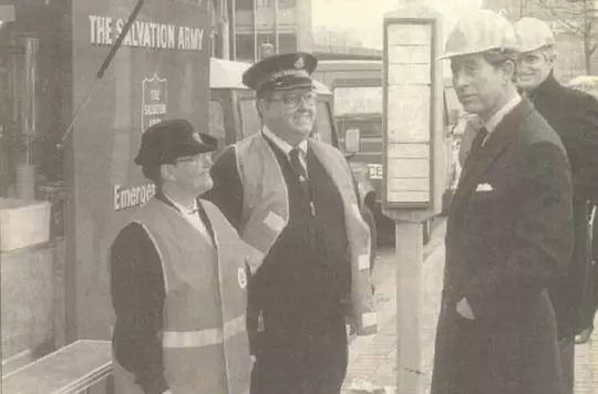 Prince Charles, on the right, speaks to two Salvation Army personnel, on the left, with a Salvation Army Emergency Response Van, a bus stop and another individual in the background.