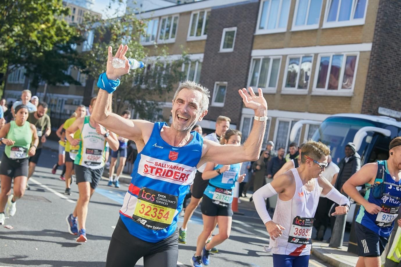 a man wearing a team sally army jersey running in the London marathon, he is smiling and waving at the camera. 