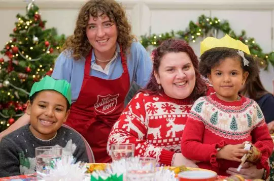 Olivia is wearing a christmas jumper and sitting at a table laid for a Christmas dinner at a Salvation Army church. Her little girl sits on her lap and her little boy on her left. They are also both wearing Christmas jumpers and hats from the crackers. A salvation Army volunteer wearing a red apron is standing with them and they are all smiling at the camera. 