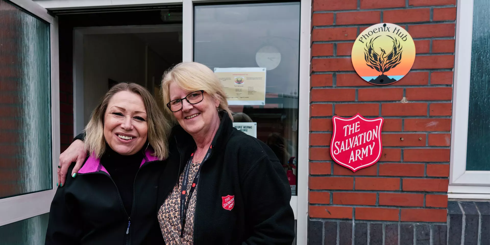 Victoria has a blonde bob and a big smile, she is standing outside her local Salvation Army with her former support worker, an older woman with short fair hair, she has her arm around Victoria's shoulder. They are both looking at the camera and smiling. 