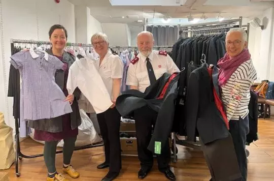 Emma Neill, Regent Hall Community Engagement Lead, Geoff and Liz Chape, Salvation Army Commanding Officers at Regent Hall, and Sonya Knight, Lead Volunteer at the Regent Hall Community Wardrobe standing in the community wardrobe and holding up a selection of school uniforms.