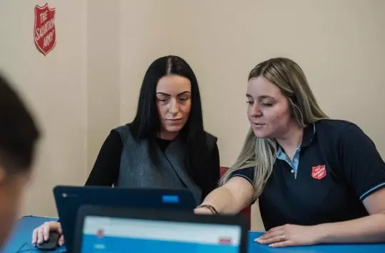 An employment plus worker with long blonde hair sits next to a young woman who is looking intently at the laptop screen as the Salvation Army worker gives her instructions. The young woman has long dark hair and is wearing a smart navy dress.