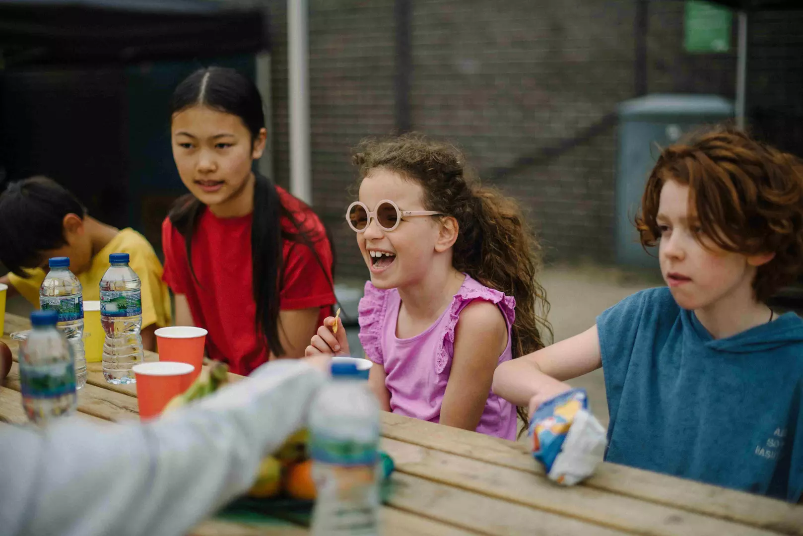 A group of primary school age children are eating luunch at a picnic table, the image is focused on one little girl with brown curly hair tied in a ponytail. She is wearing a purple t-shirt and pink heart shaped glasses. She's laughing and looking at someone off camera. 