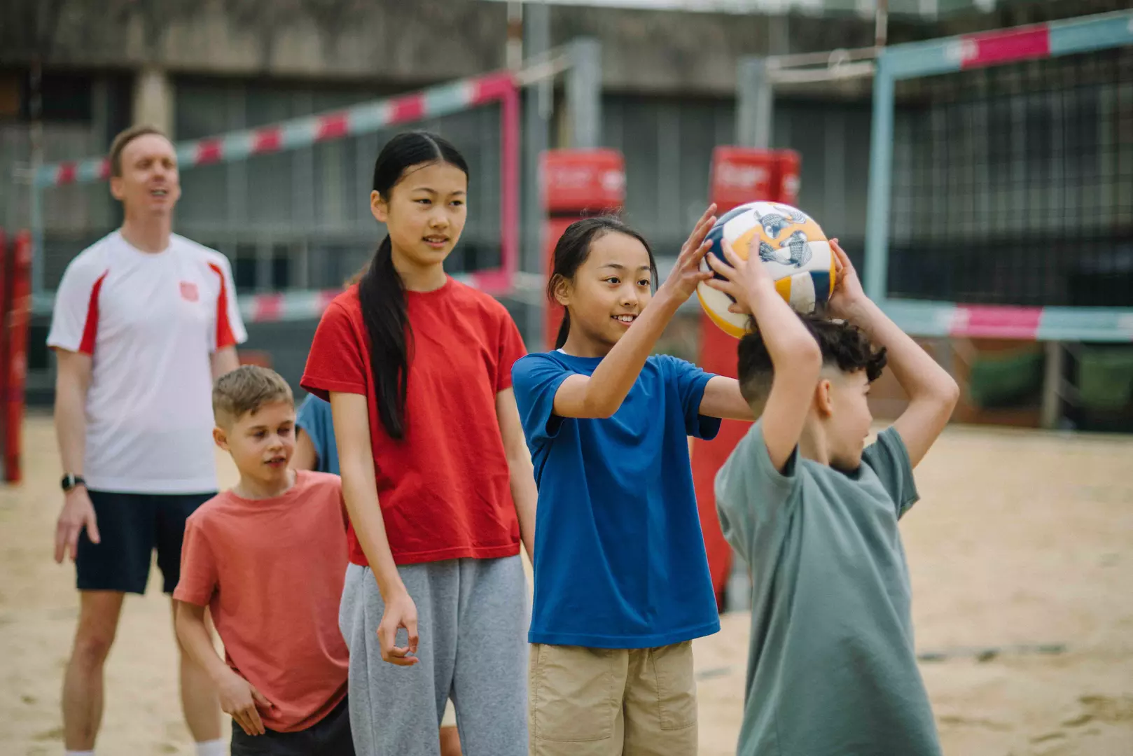 A group of primary school aged kids wearing bright summer clothes are playnig with a football at a summer camp as a Salvation Army officer in a white polo shirt cheers them on.