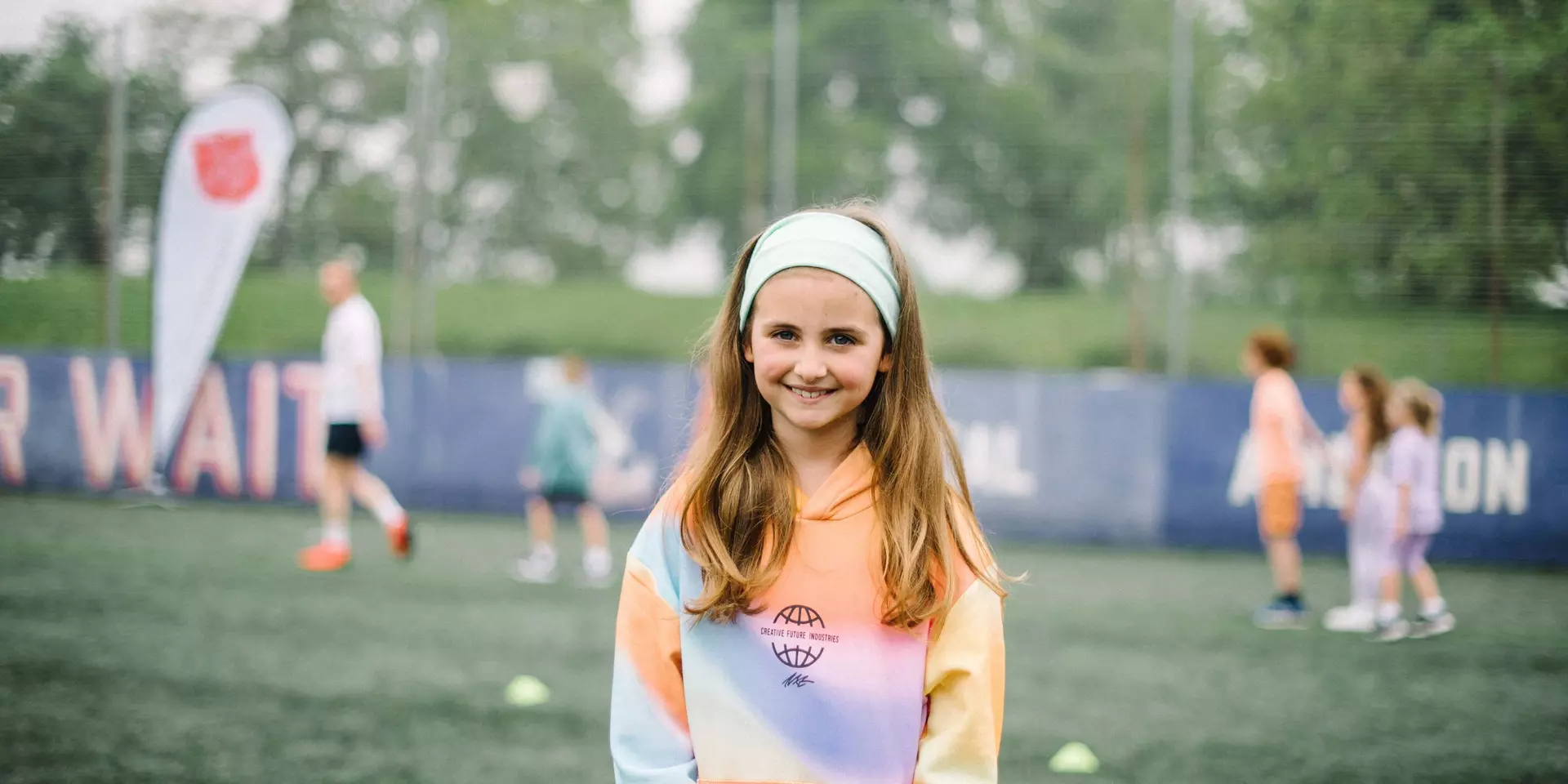 A young girl with a colourful hoodie and a headband, standing on artificial grass with people playing football in the background.