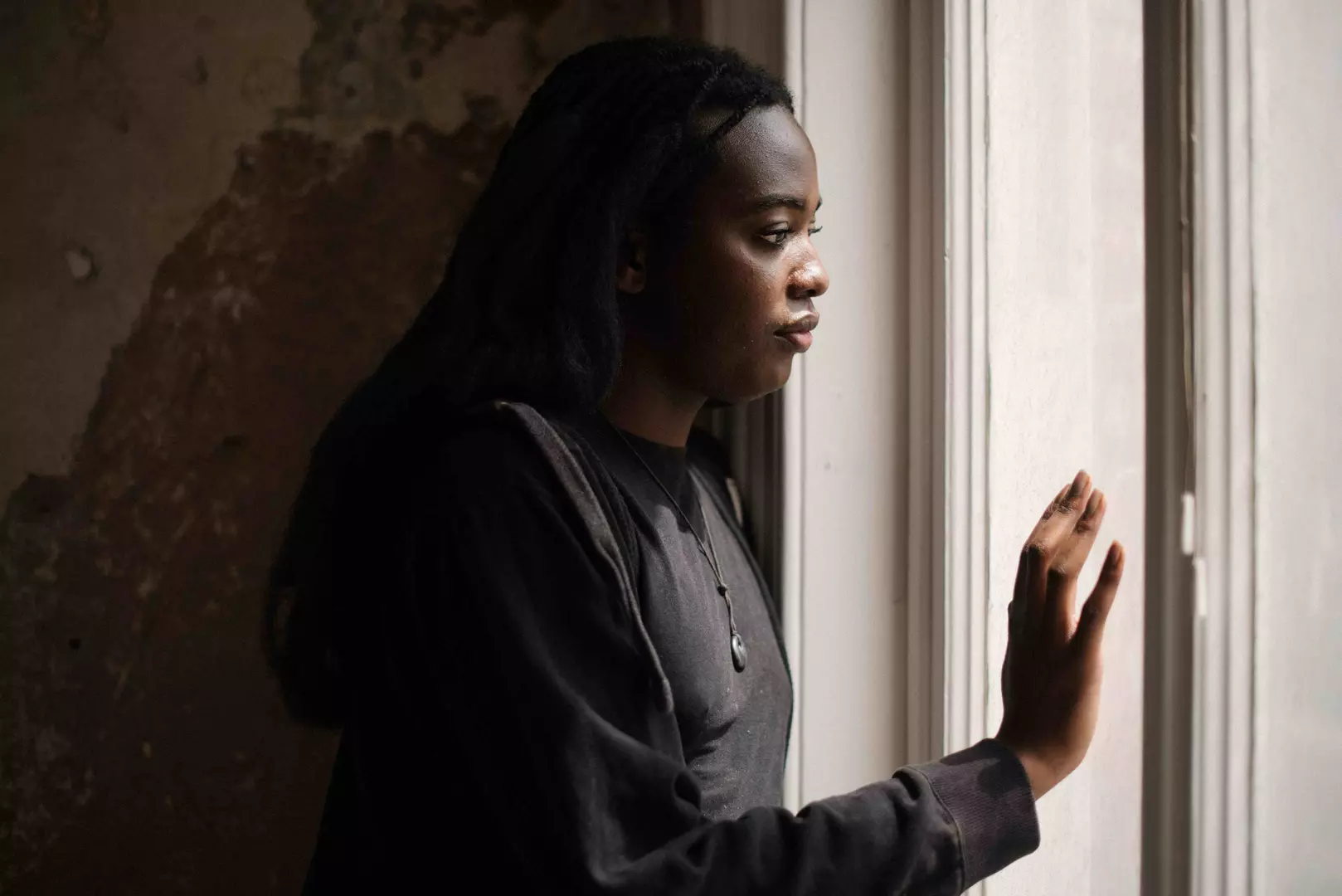 Rose, a young black woman with long dark hair, stands looking out of a window. She holds her hand up to the light. 